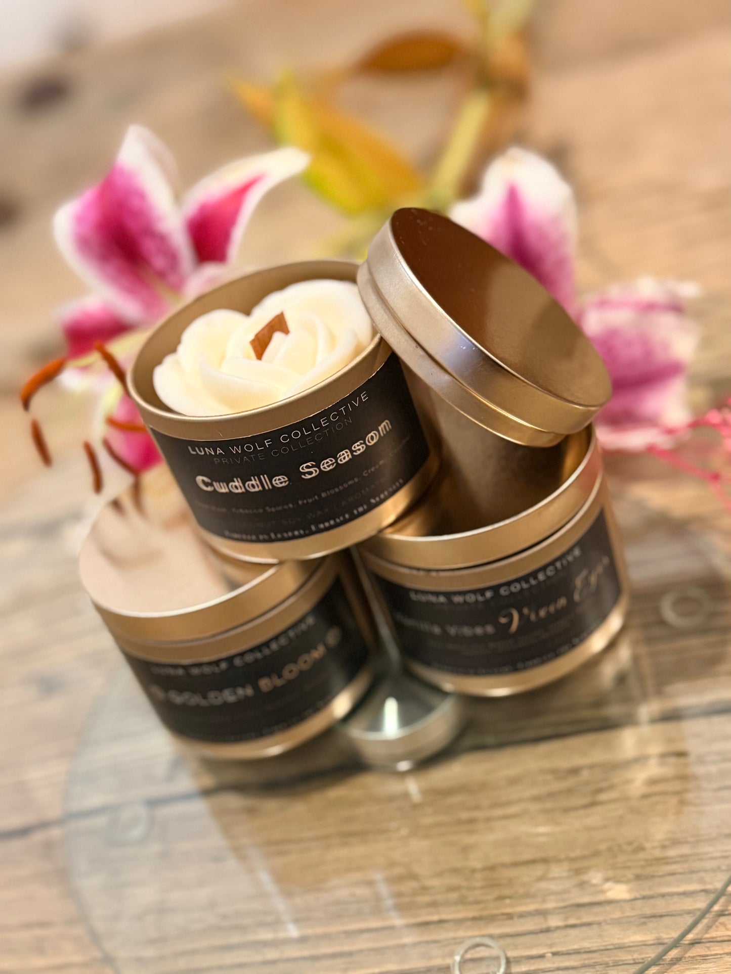 Three candles in metallic tins with visible labels on a wooden surface with flowers in the background