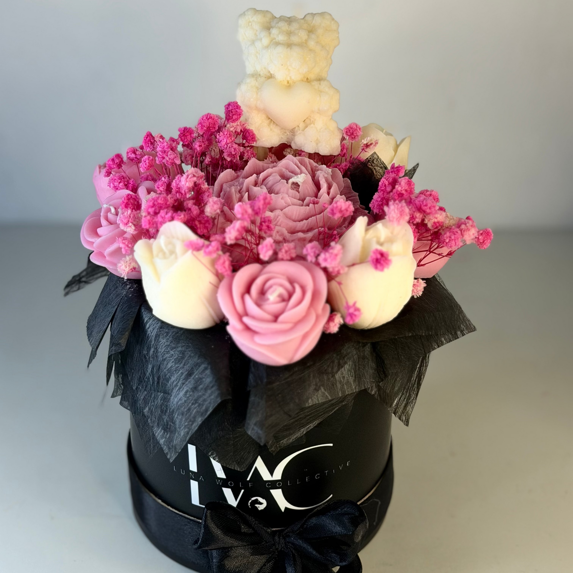 A round black gift box with a black ribbon bow, decorated with pink and cream artificial flowers and a small cream teddy bear on top. The front of the box features the white printed logo 'LWC COLLECTIONS'.